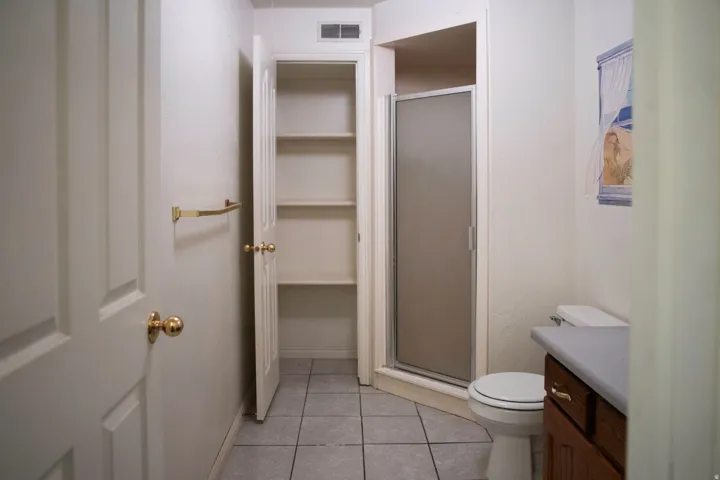 Bathroom featuring a corner shower with a frosted glass door, a built-in linen closet with shelving, a vanity with a dark wood-finish cabinet and a light-colored countertop, and square tile flooring