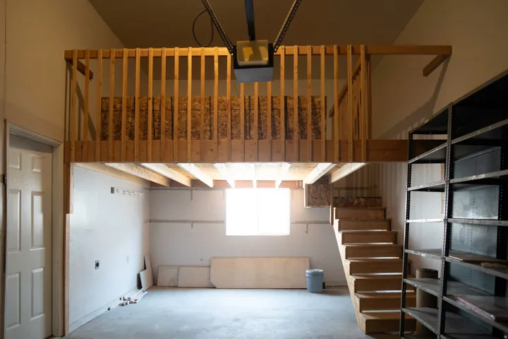 Spacious garage featuring a built-in wooden loft with railing, wooden stairs, a single window, and a white interior door