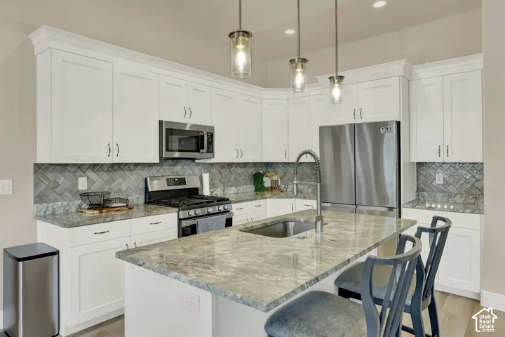 Spacious kitchen featuring an island with an undermount sink, white cabinetry with crown molding, stainless steel appliances, a gray herringbone tile backsplash, and light-toned countertops