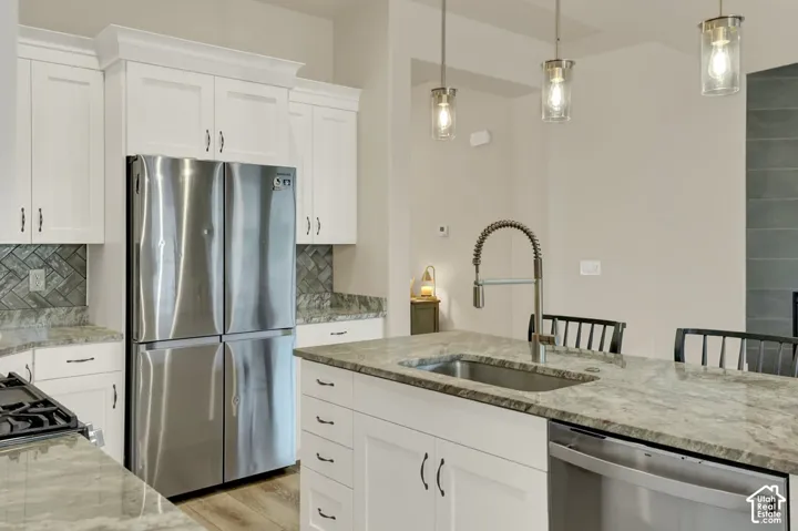 Kitchen featuring white cabinetry, stainless steel appliances, herringbone tile backsplash, an island with an undermount sink, and pendant lighting