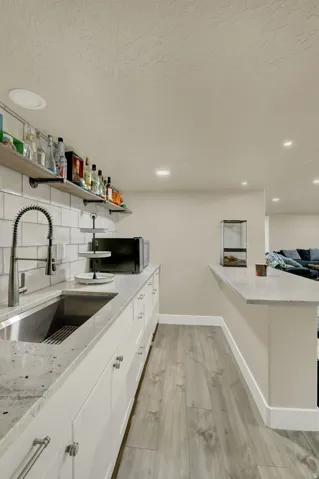 Wet bar featuring a stainless steel sink, granite-style countertop, white shaker-style cabinetry, subway tile backsplash, and open shelving