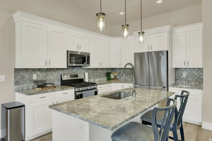 Spacious kitchen featuring an island with an undermount sink, white cabinetry with crown molding, stainless steel appliances, a gray herringbone tile backsplash, and light-toned countertops