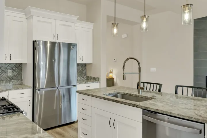 Kitchen featuring white cabinetry, stainless steel appliances, herringbone tile backsplash, an island with an undermount sink, and pendant lighting