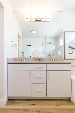 Dual vanity with light gray cabinetry, brushed gold hardware, gray countertops, and wood-finish flooring