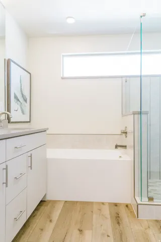 Modern bathroom featuring a built-in soaking tub with tile surround, a glass-enclosed shower, a vanity with white cabinetry, and wood-finish flooring