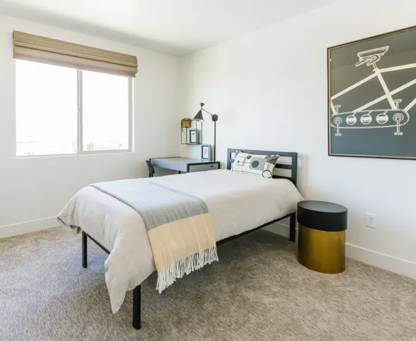 Light-filled bedroom featuring a large window with privacy shade, neutral-toned carpet flooring, white baseboards, and a dedicated desk area with shelving and task lighting