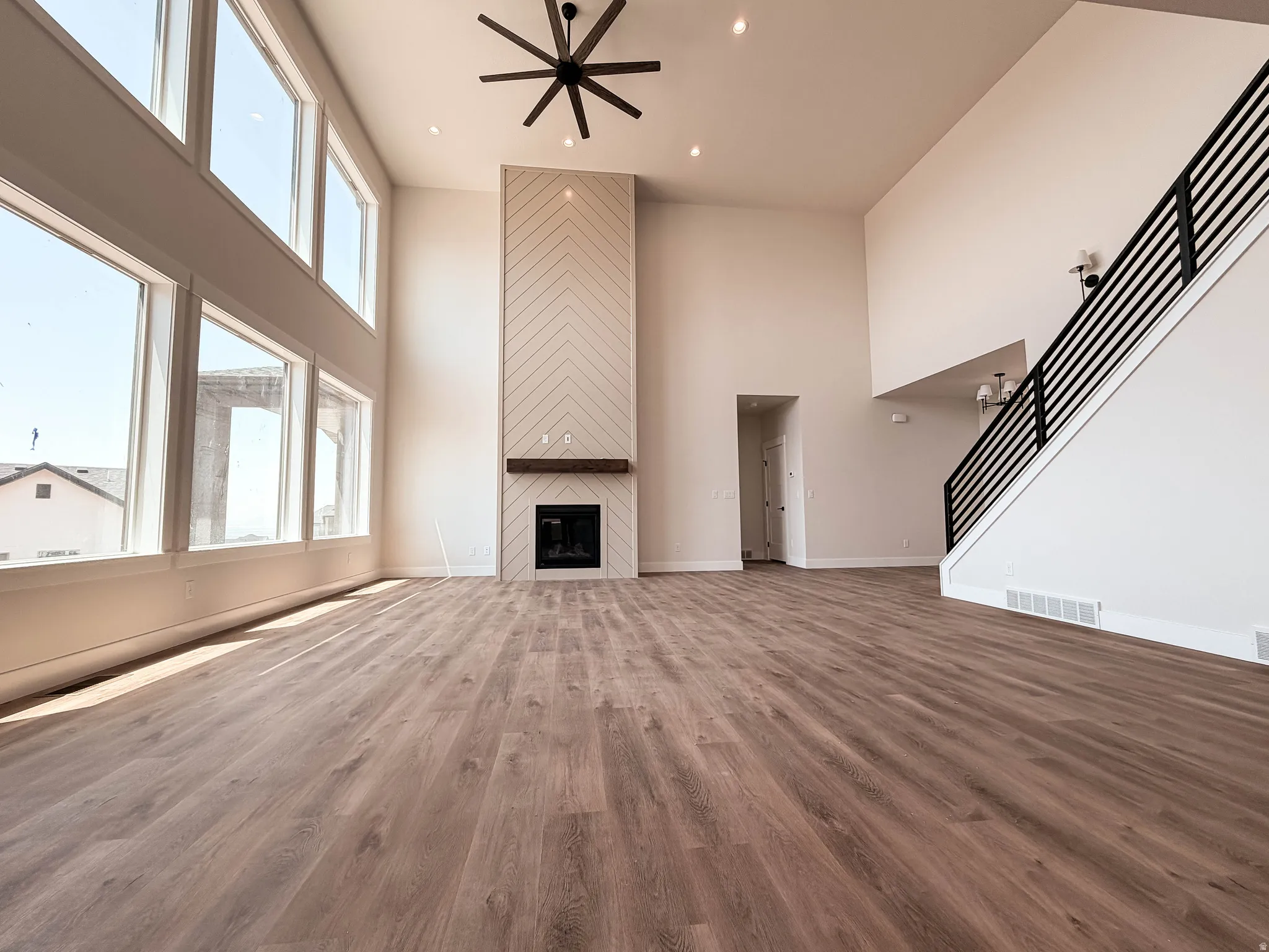 Spacious living area featuring double-height ceilings, a floor-to-ceiling fireplace with a chevron-patterned facade and wood mantel, large windows, wood-finish flooring, and a modern ceiling fan