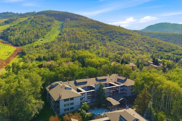 Multi-story building surrounded by dense mature trees, featuring a visible outdoor pool