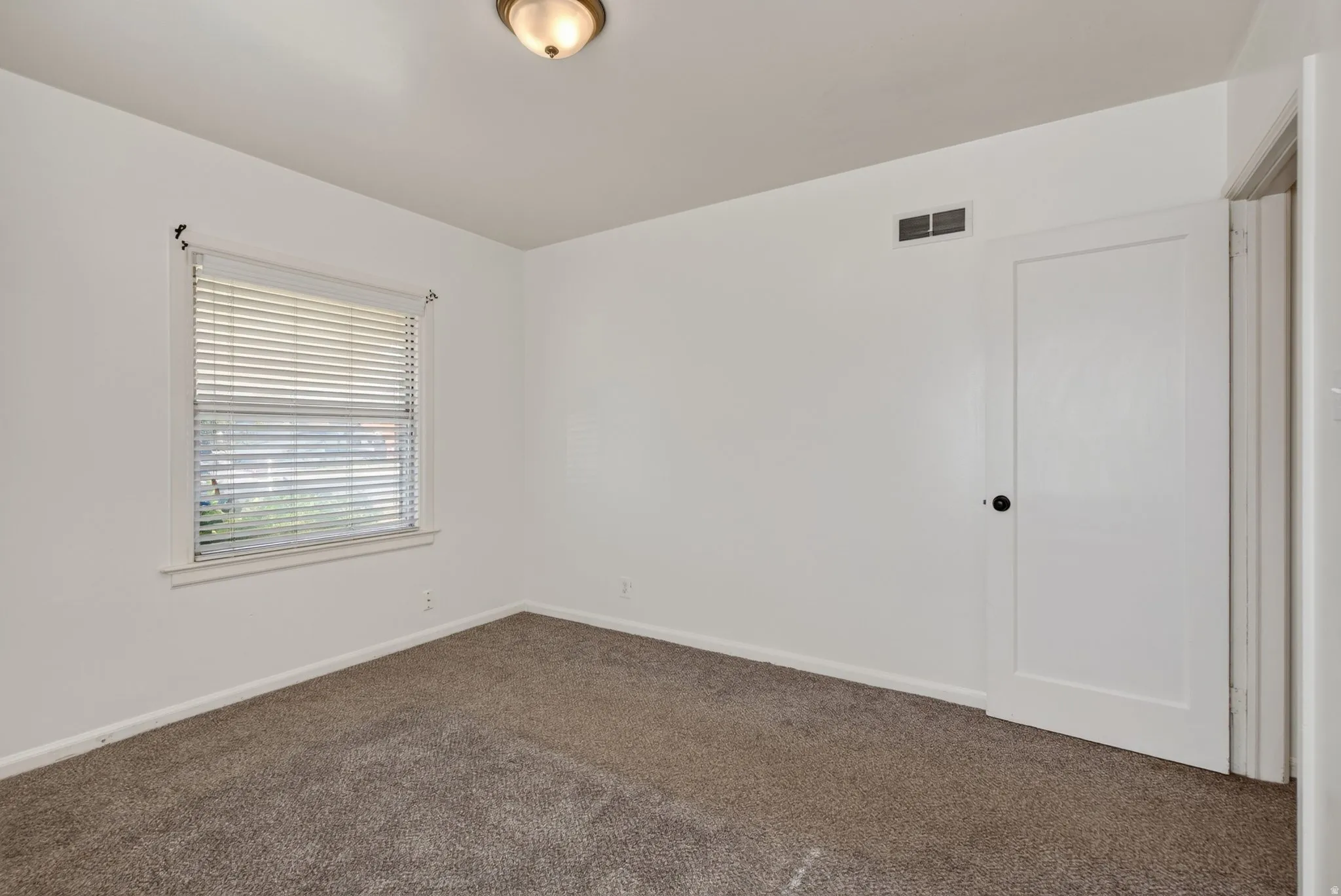 Bedroom featuring neutral-toned carpeting, white walls, a single window with blinds, a white door with dark hardware, and a ceiling-mounted light fixture
