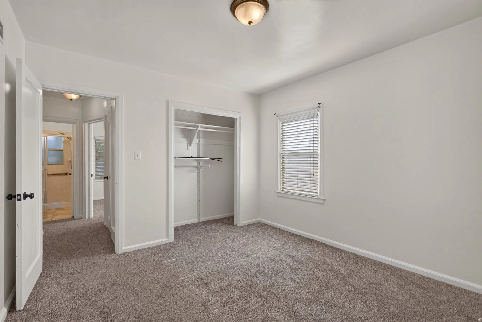 Neutral-toned wall paint and carpeted flooring in a bedroom featuring a window with blinds, a built-in closet with shelving and hanging rods, and a ceiling-mounted light fixture