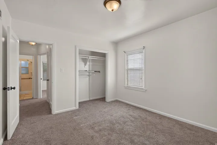 Neutral-toned wall paint and carpeted flooring in a bedroom featuring a window with blinds, a built-in closet with shelving and hanging rods, and a ceiling-mounted light fixture