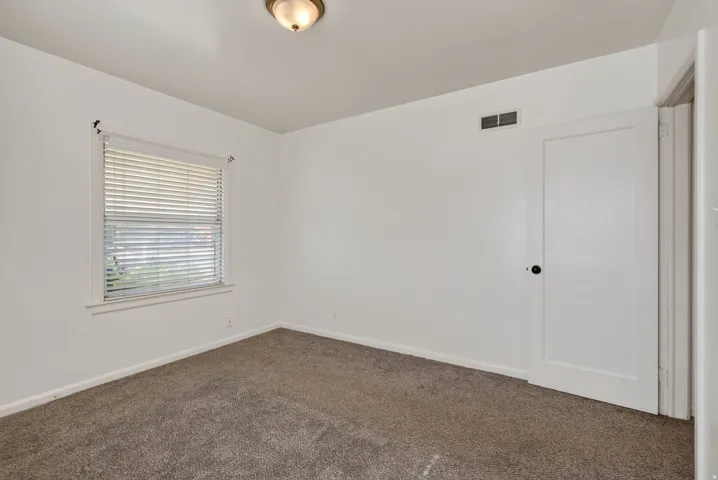 Bedroom featuring neutral-toned carpeting, white walls, a single window with blinds, a white door with dark hardware, and a ceiling-mounted light fixture