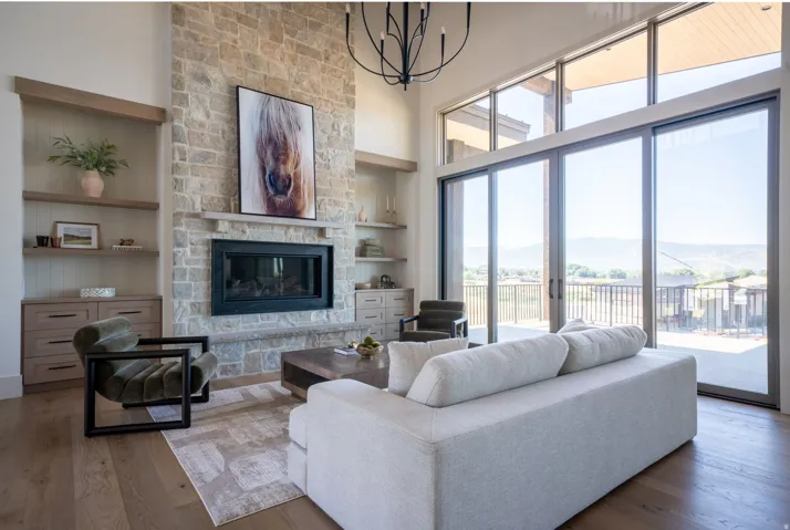 Soaring ceilings with extensive window wall, stone-clad fireplace, integrated shelving, and wood-finish flooring