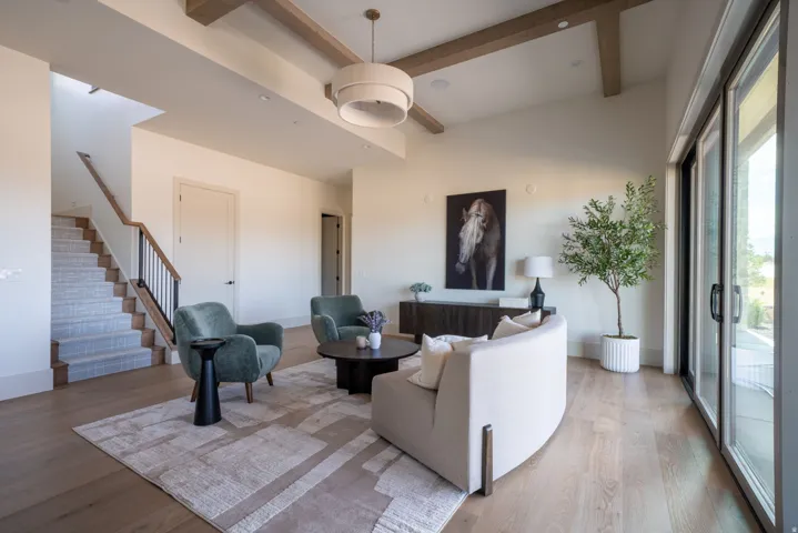 High ceilings with exposed wood beams, light wood-finish flooring, staircase with carpeted treads and metal railing, built-in shelving, and large sliding glass doors