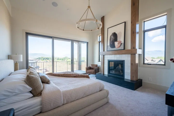 Sliding glass doors to an exterior balcony, light-toned tiled fireplace with a wood-beam mantel, wood-finish structural columns, and a contemporary chandelier