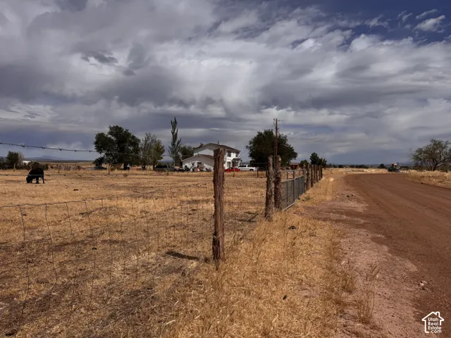 Rural setting featuring a dirt road, wire fencing, mature trees, and a distant two-story residence