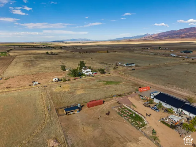 Aerial acreage featuring expansive open terrain, a multi-structure property layout, raised-bed garden plots, and a prominent mountain range backdrop