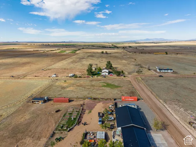 Aerial view of expansive acreage featuring a large metal-roofed structure, fenced garden plots, and distant mountain terrain