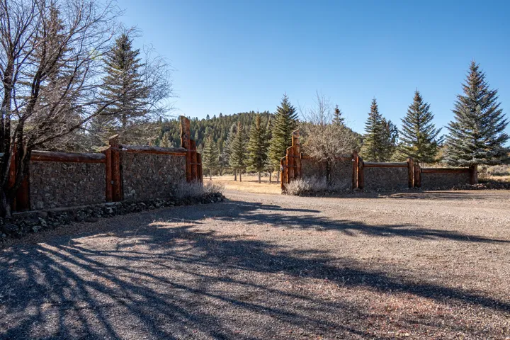 Stone and wood-finish entry wall with a gravel driveway, surrounded by mature evergreen trees