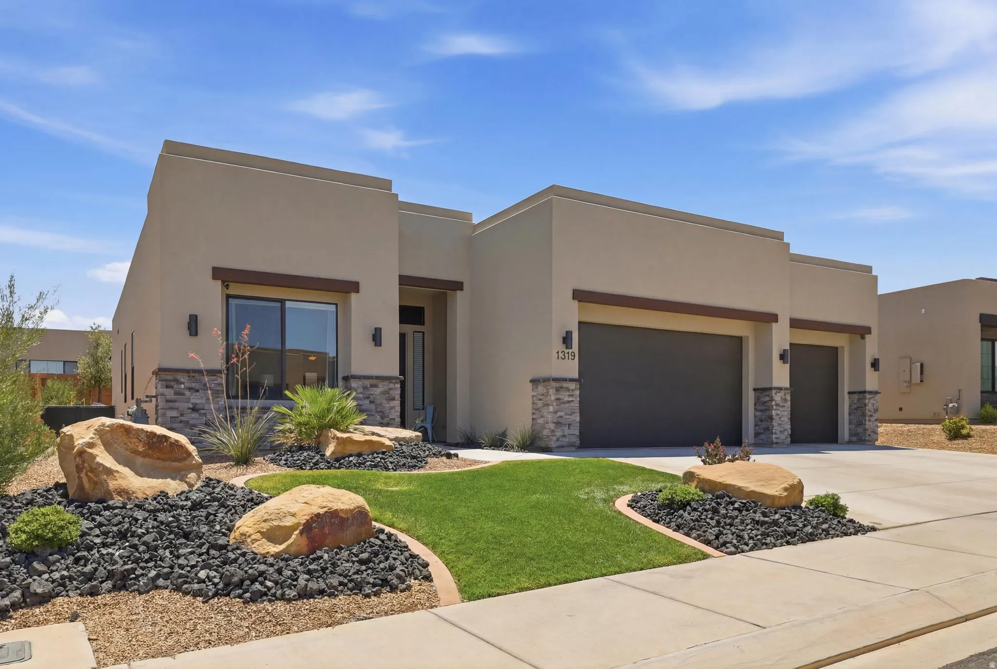 Contemporary stucco exterior featuring stone accents, a dual garage, desert landscaping with large rock features, a manicured lawn, and a concrete driveway