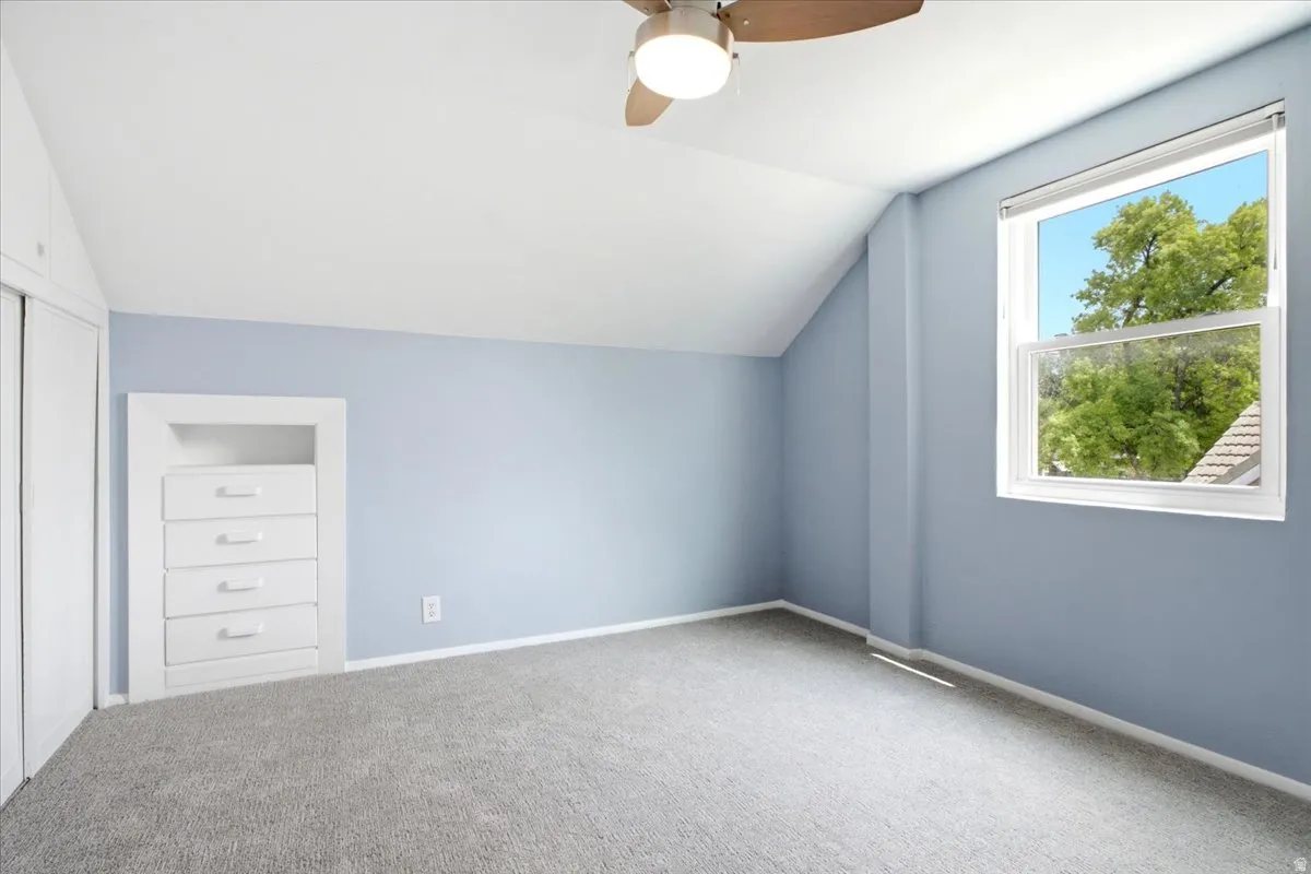Carpeted room featuring an angled ceiling, a ceiling fan with light, a window with white trim, and a built-in dresser with four drawers