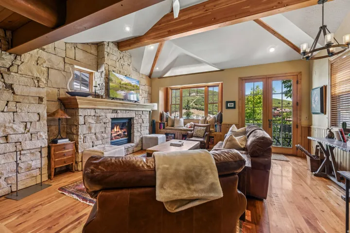 Living area featuring a stone fireplace with a wood mantle, wood-finish flooring, vaulted ceilings with exposed wood beams, and French doors opening to an exterior setting