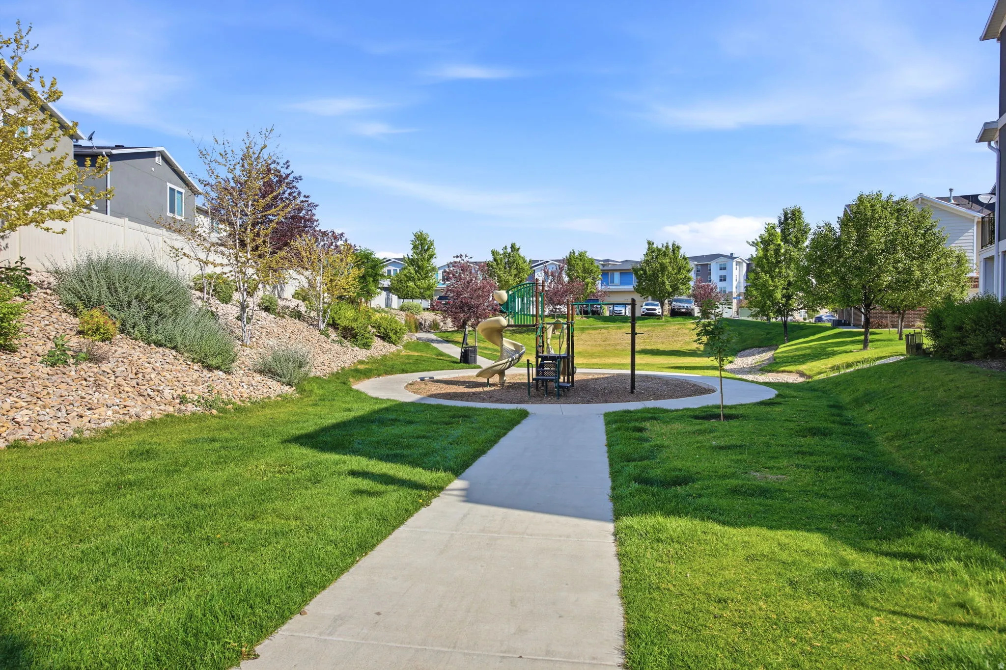 Community green space featuring a playground area with a slide and climbing structure, surrounded by mature trees and manicured lawns