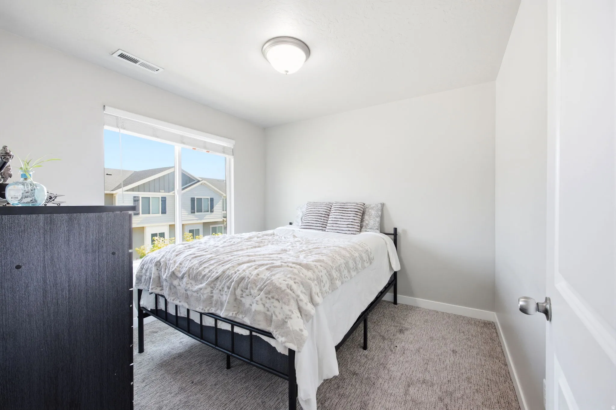 Bright bedroom featuring a large window with roller shades, light gray wall paint, and gray carpet flooring