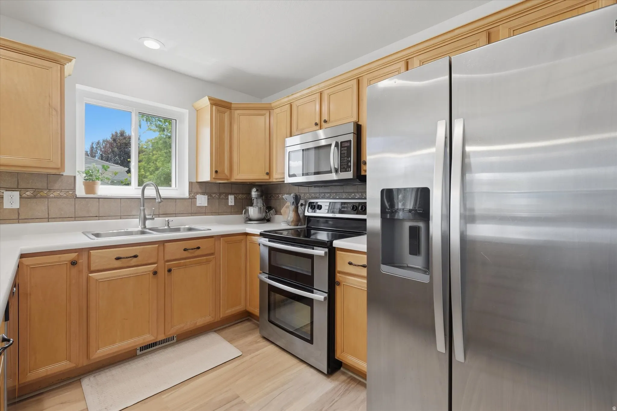 Kitchen featuring light wood cabinetry, stainless steel appliances, a double basin sink with a high-arc faucet, white countertops, and a tiled backsplash
