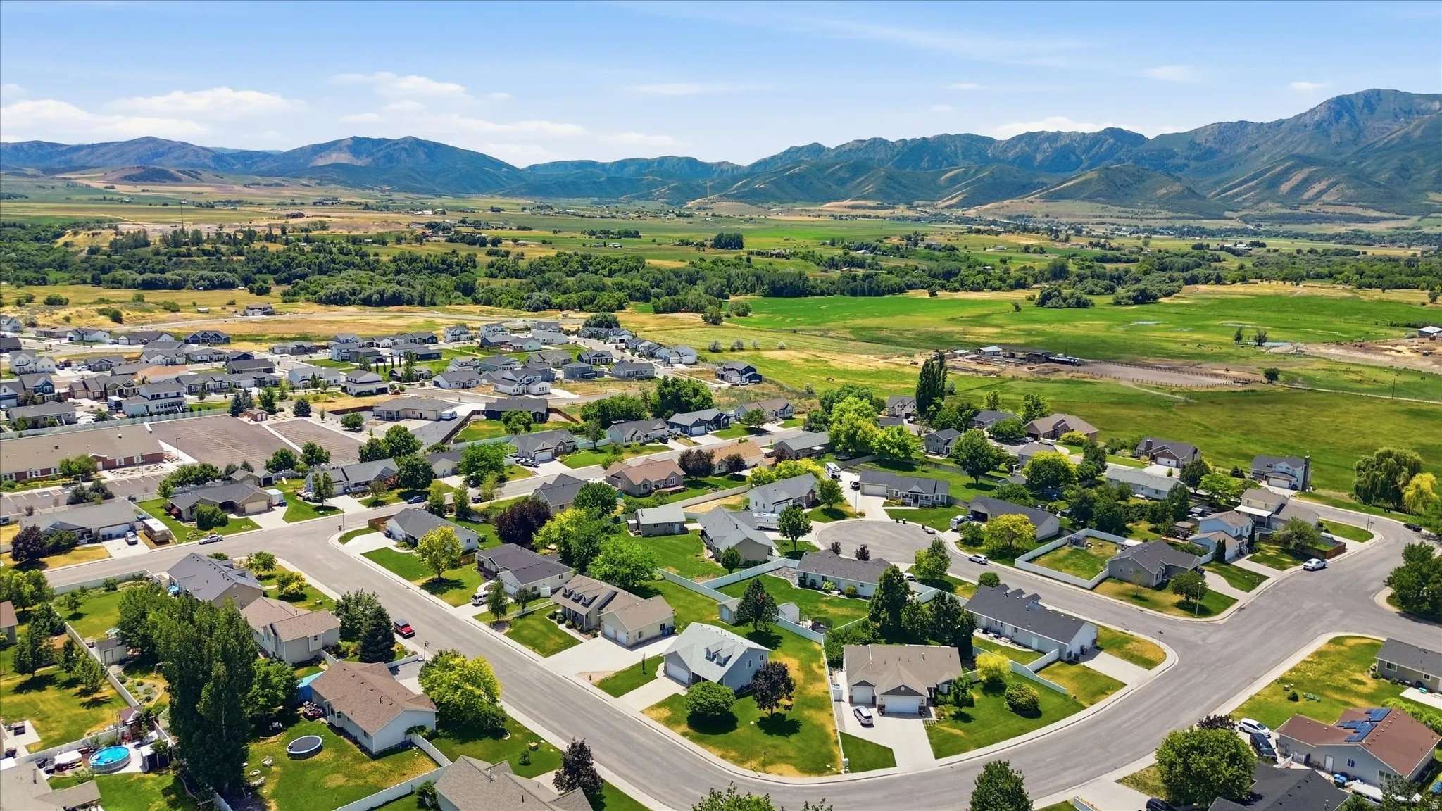 Residential community featuring paved streets, mature landscaping, and single-story homes, set against a backdrop of distant mountains and expansive green fields