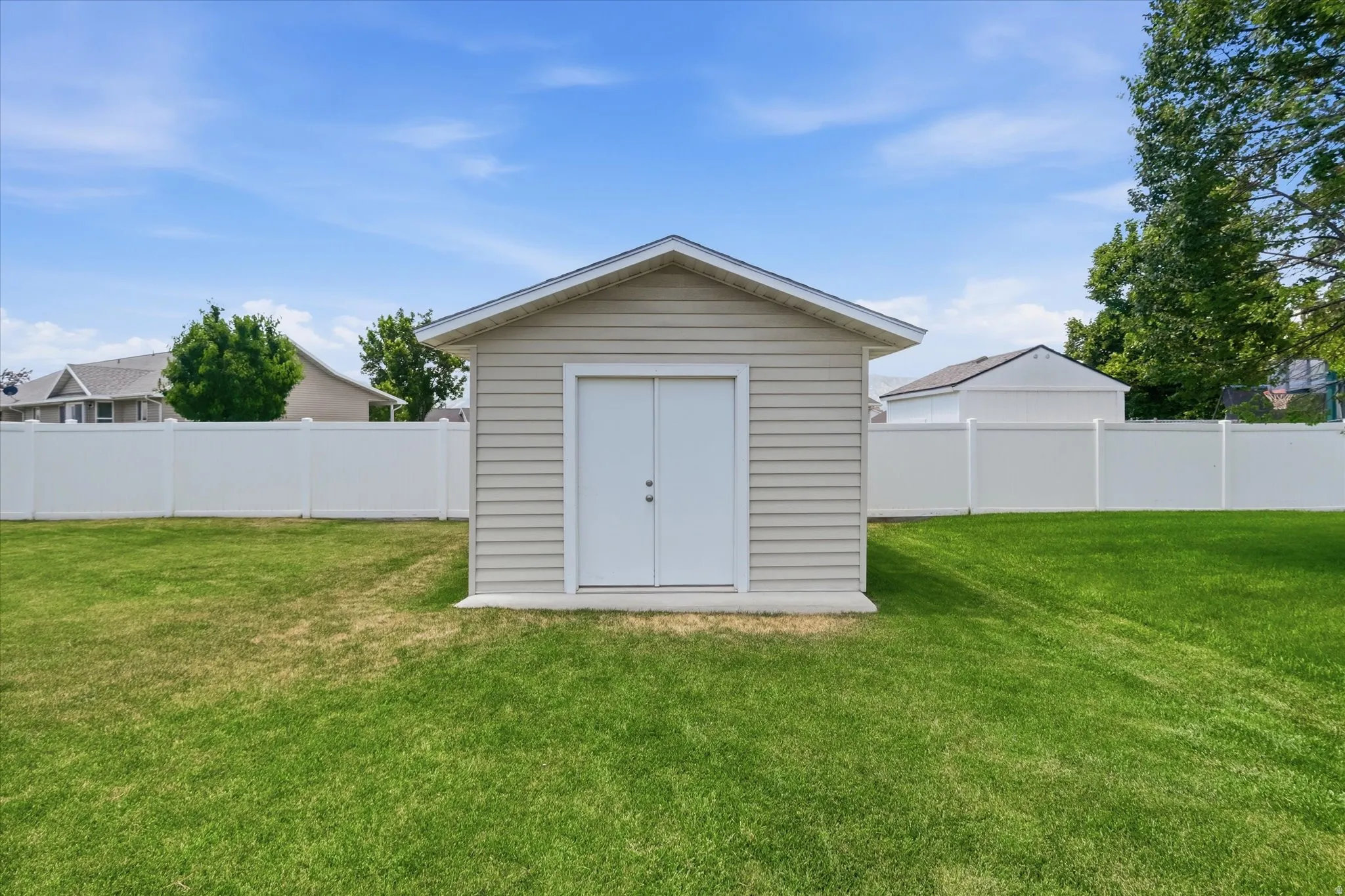 Detached storage shed with siding exterior, dual entry doors, and a partial concrete foundation, set within a spacious grassy yard featuring a white privacy fence