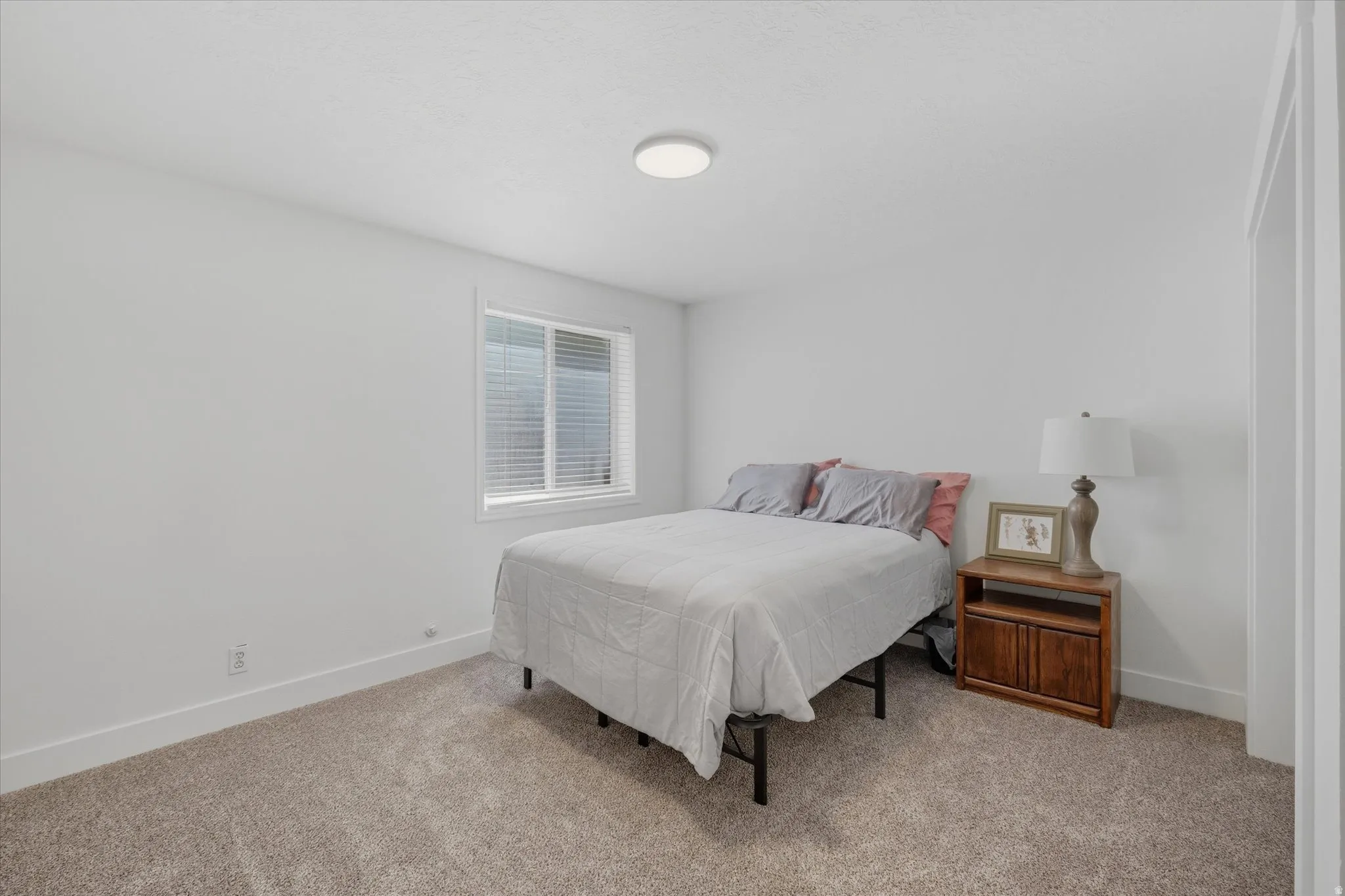 Bedroom featuring light-colored carpeting, white walls, a single window with horizontal blinds, a white ceiling, and a flush-mount circular light fixture