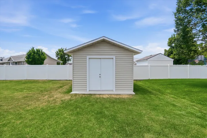 Detached storage shed with siding exterior, dual entry doors, and a partial concrete foundation, set within a spacious grassy yard featuring a white privacy fence
