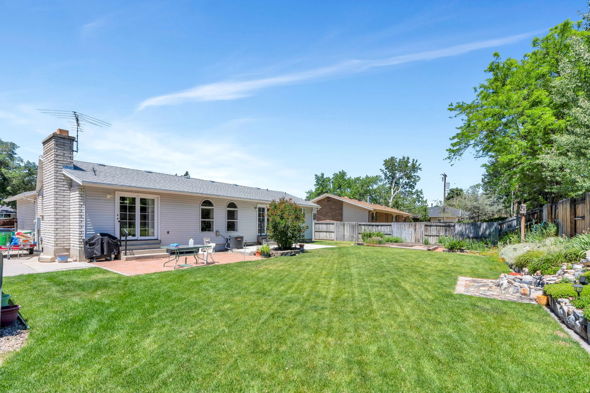 Expansive backyard featuring a large green lawn, a brick paver patio, a decorative rock garden, and mature perimeter trees