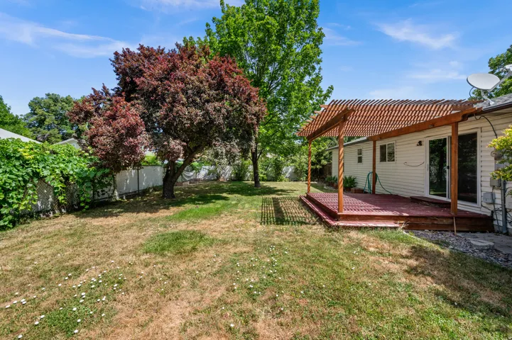 Expansive backyard featuring a wood deck with a slatted pergola, sliding glass door access to the interior, mature trees providing natural shade, and perimeter fencing