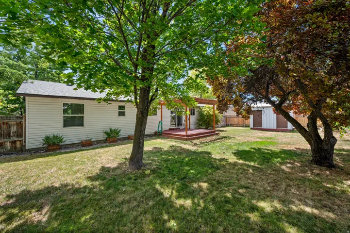 Expansive backyard featuring a red wood-finish deck with a pergola, white siding exterior, mature trees, and a detached shed