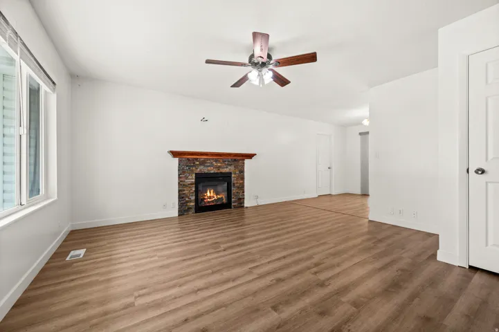 Living area featuring a stone-faced fireplace with a wood mantel, wood-finish flooring, a ceiling fan, and large windows with blinds