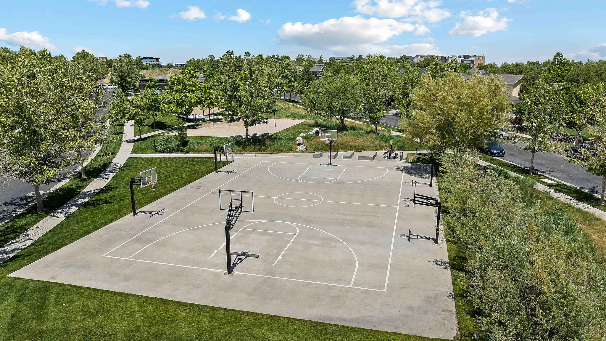 Concrete basketball court featuring multiple hoops, surrounded by landscaped green spaces and mature trees