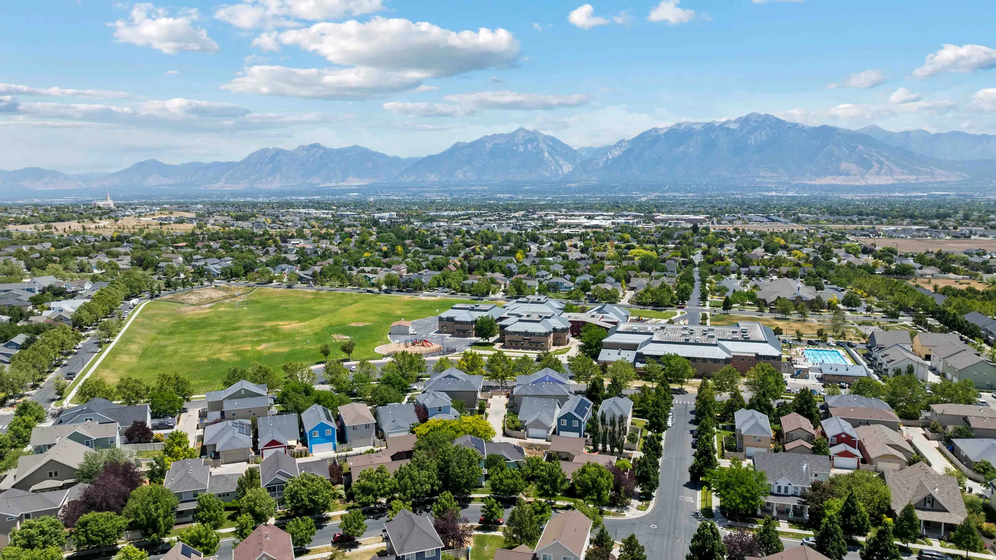 Expansive aerial capturing a residential area with diverse home styles, a large green field, and distant mountains, community center & pool
