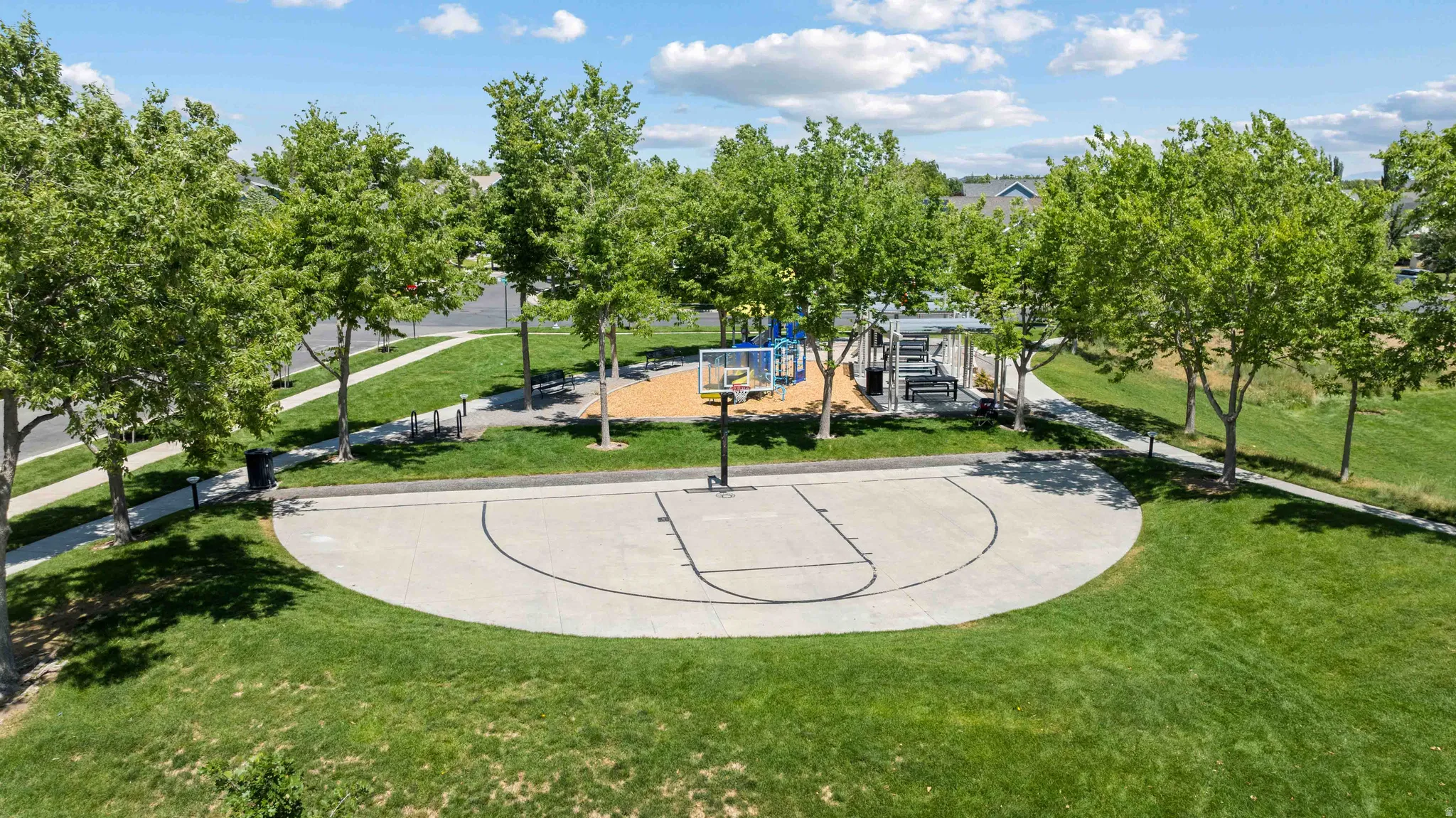 Expansive green space featuring a concrete half-basketball court, shaded gathering area with benches, and a playground structure