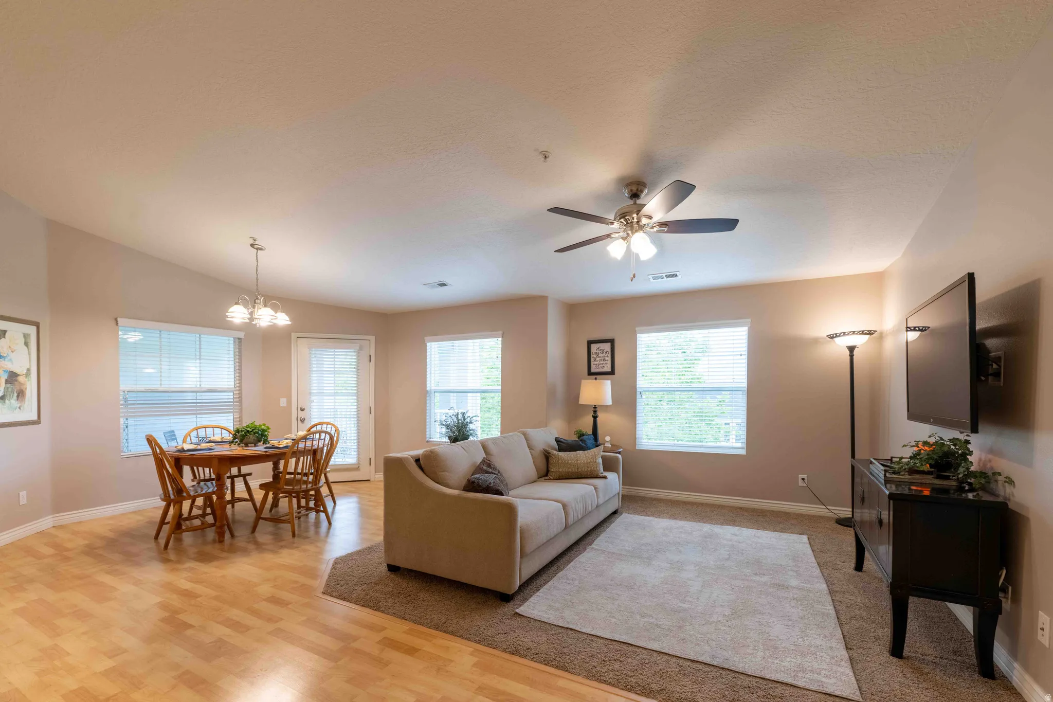 Open-concept living space featuring wood-finish flooring, a ceiling fan with integrated lighting, multiple windows with blinds, and a glass-paneled door
