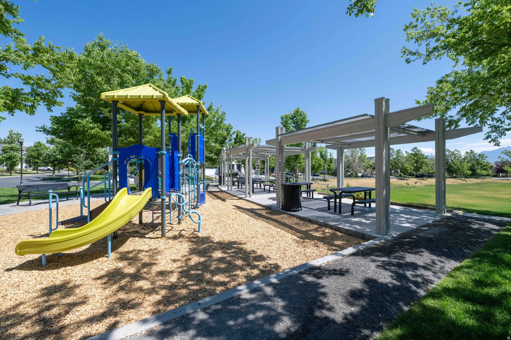 Outdoor recreation area featuring a vibrant playground with wood chip surfacing, a multi-section picnic pergola, and paved walking paths