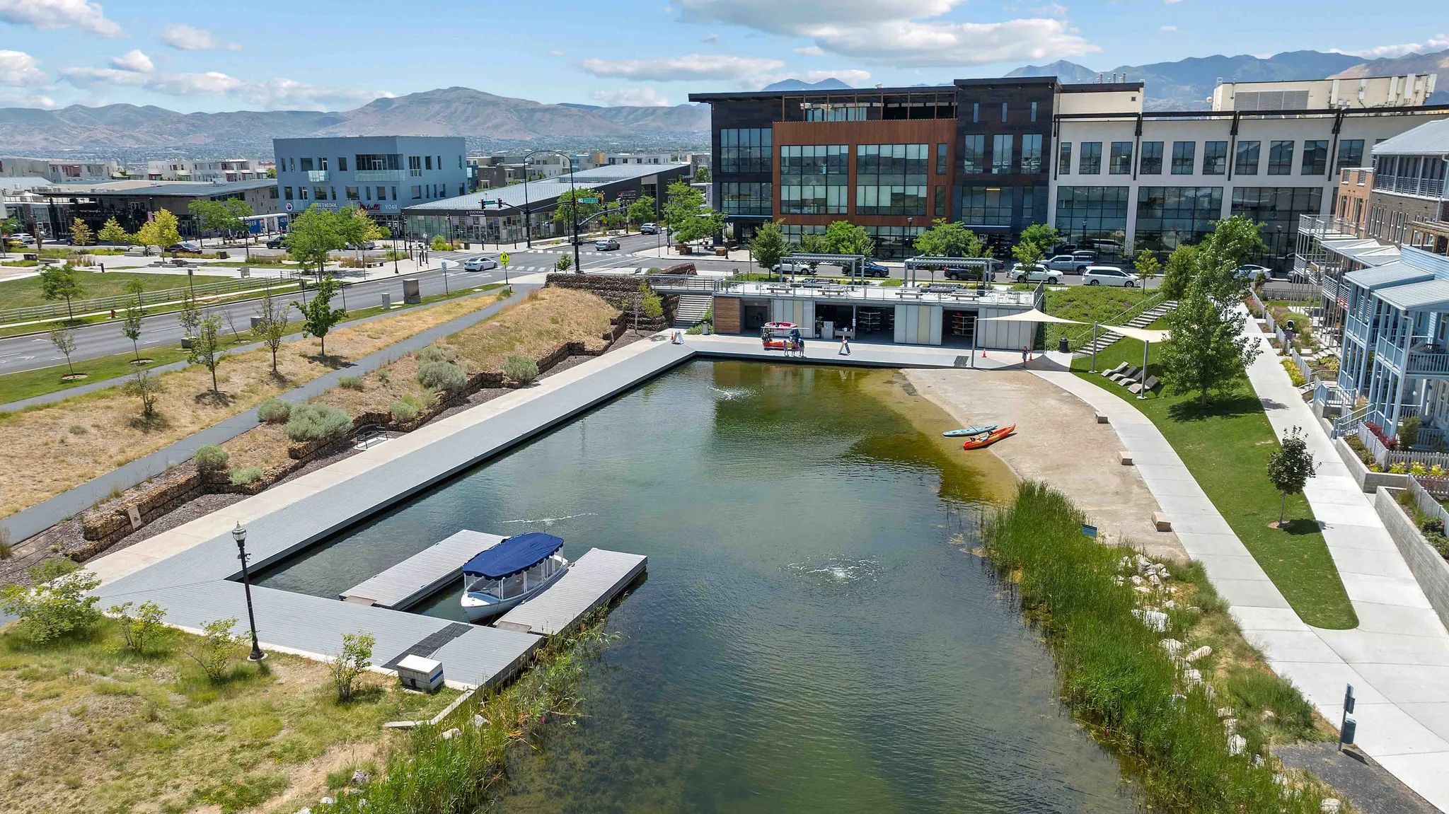 Waterfront property featuring a private boat dock, sandy beach area, established pedestrian paths, modern commercial structures, and distant mountain scenery