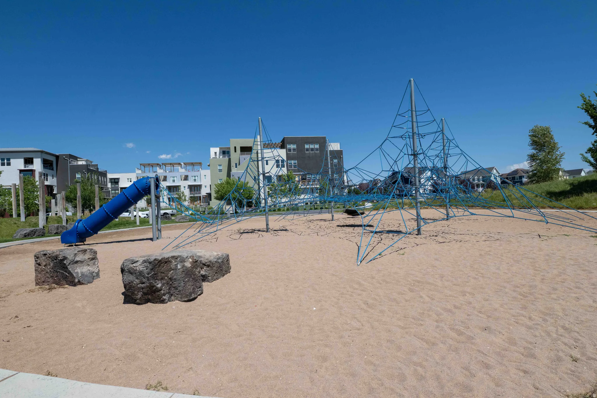Community park featuring a blue tube slide, a rope climbing structure, and natural rock elements on a sand-covered ground