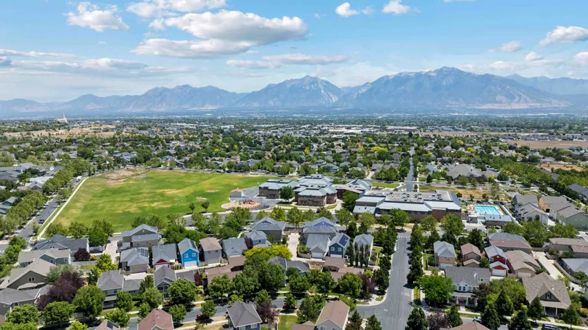 Expansive aerial capturing a residential area with diverse home styles, a large green field, and distant mountains, community center & pool