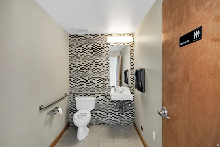Restroom featuring a wall-mounted toilet, pedestal sink, mosaic tile accent wall, and a full-width mirror with overhead lighting