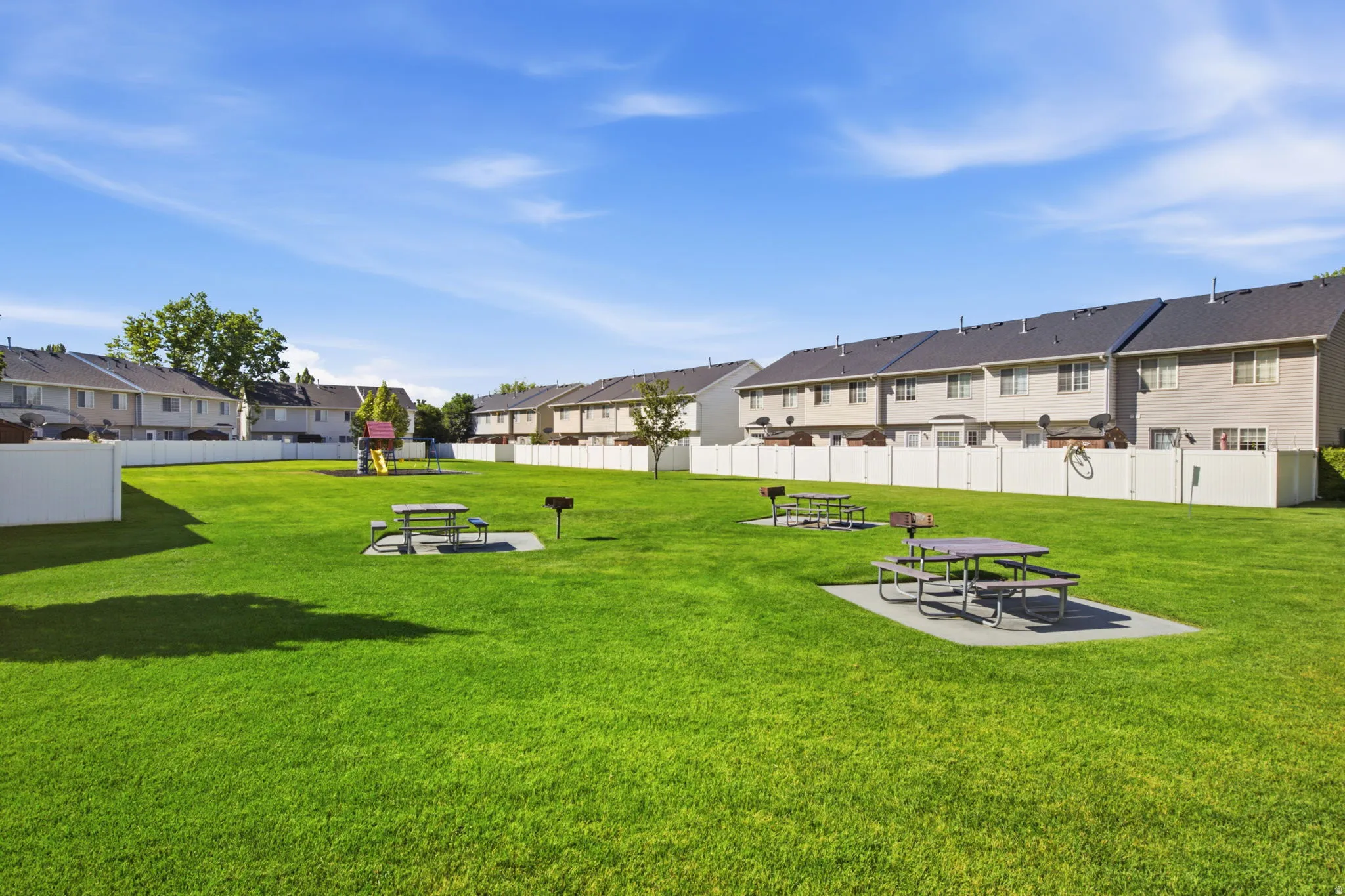 Expansive community green space featuring a manicured lawn, multiple picnic tables on concrete pads, built-in barbecue grills, and a dedicated play structure