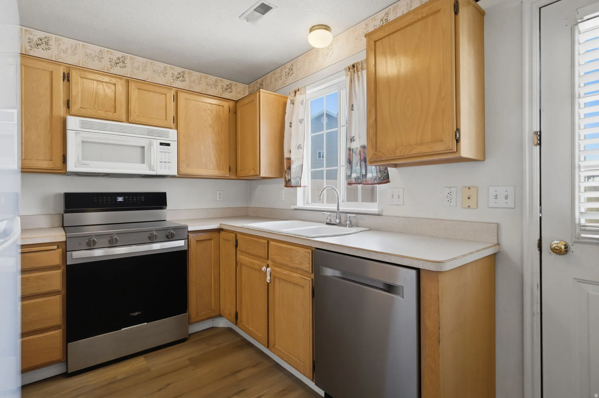 Kitchen featuring light wood cabinetry, a stainless steel range, a built-in microwave, a white double-basin sink with a chrome faucet, and wood-finish flooring
