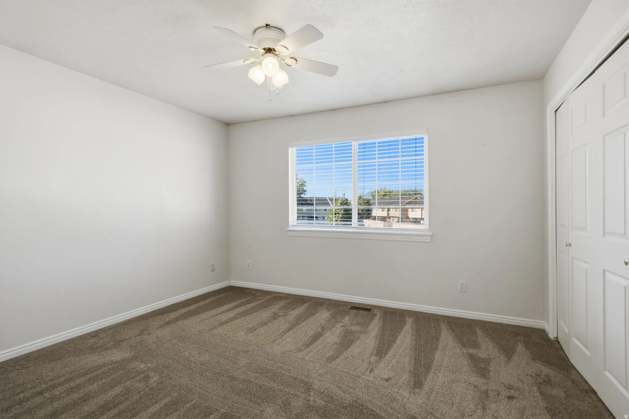 Spacious room featuring neutral carpeting, a large window, white trim, a ceiling fan with light fixture, and a bi-fold closet door