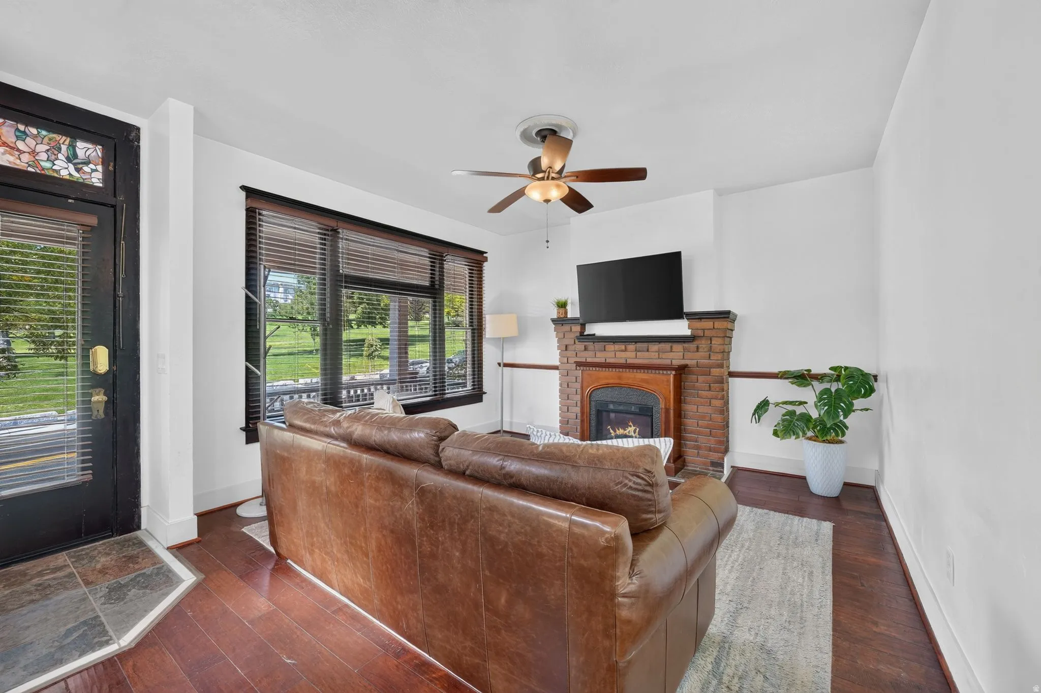 Living area featuring laminate flooring, a brick fireplace with a wood mantel, a ceiling fan with light fixture, and multiple windows